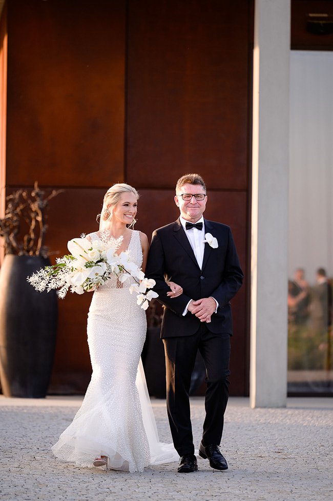 The bride, holding a bouquet of white flowers, walks arm in arm with her father, both smiling as they enter the wedding venue.