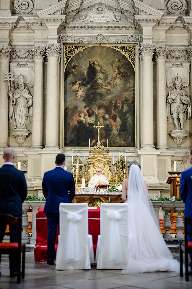 A bride and groom stand before an officiant in a beautifully decorated church during their wedding ceremony at Valtice.