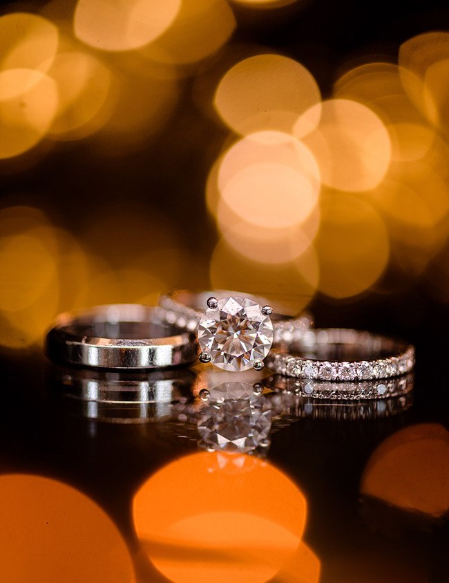 Close-up of wedding rings with a sparkling diamond engagement ring, set against a blurred background of golden lights.