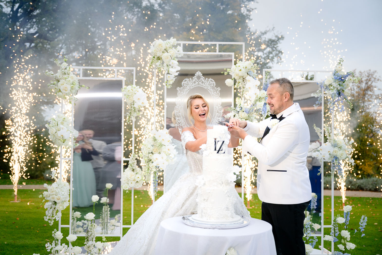 Bride and groom cutting wedding cake under a decorative arch with sparklers at Chateau Mcely, surrounded by elegant floral arrangements.