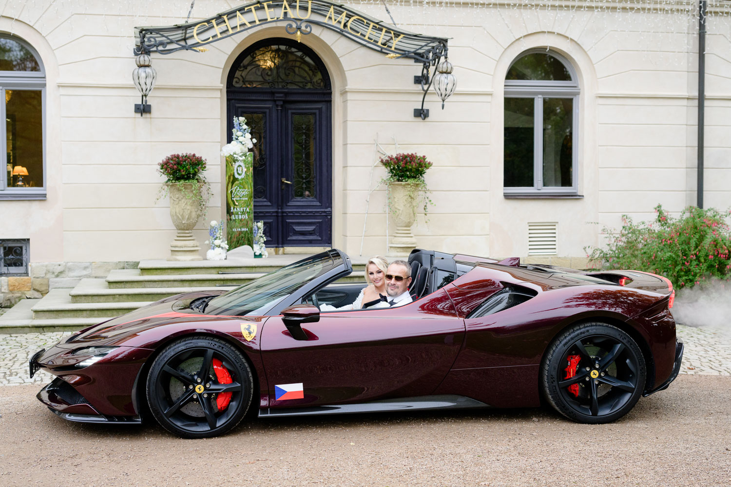 A couple in a red convertible sports car posing at the entrance of Chateau Mcely, with decorative plants and the chateau's facade in the background.