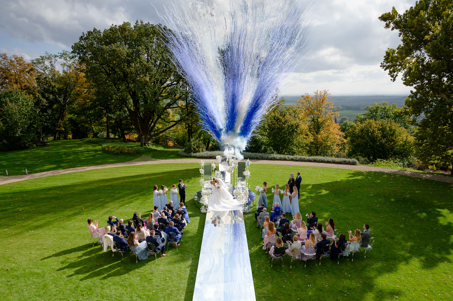 Outdoor wedding ceremony at Chateau Mcely with guests seated on a lush green lawn, a white aisle, and a dramatic blue and white confetti display.