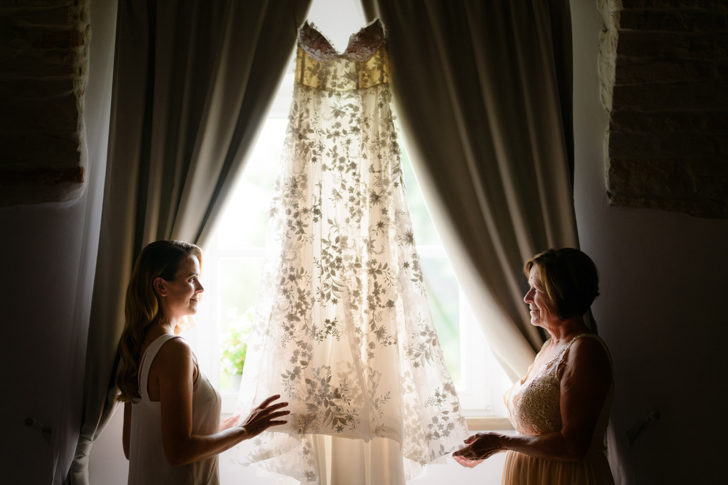A bride and her mother admiring a beautiful white lace wedding dress hanging in front of a sunlit window at na Kmine
