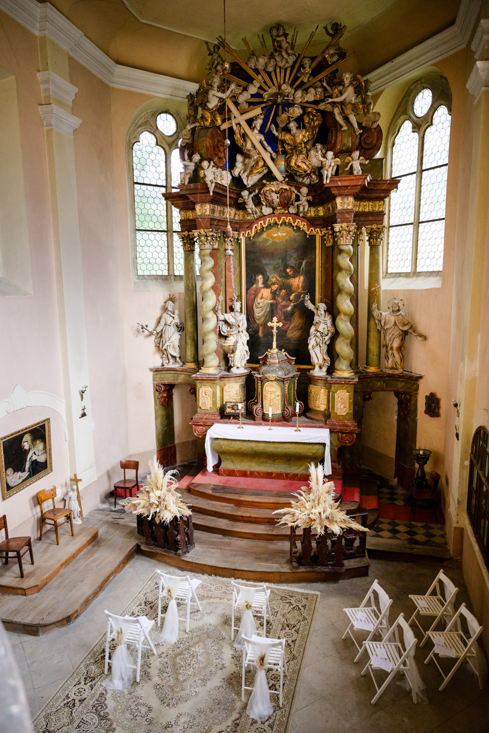 Baroque church ceremony setup with white chairs and pampas grass floral decor at Na Kmine wedding venue – captured by Jindrich Nejedly Photography
