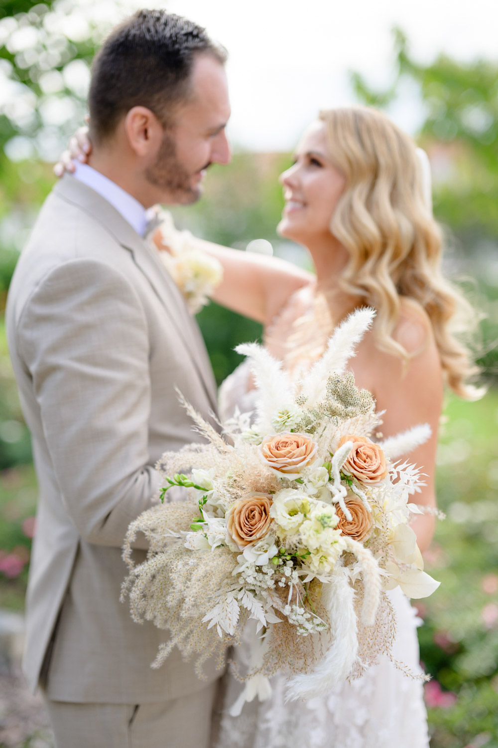 A bride and groom smiling at each other, with the bride holding a bouquet of peach and white flowers, set against a lush garden backdrop