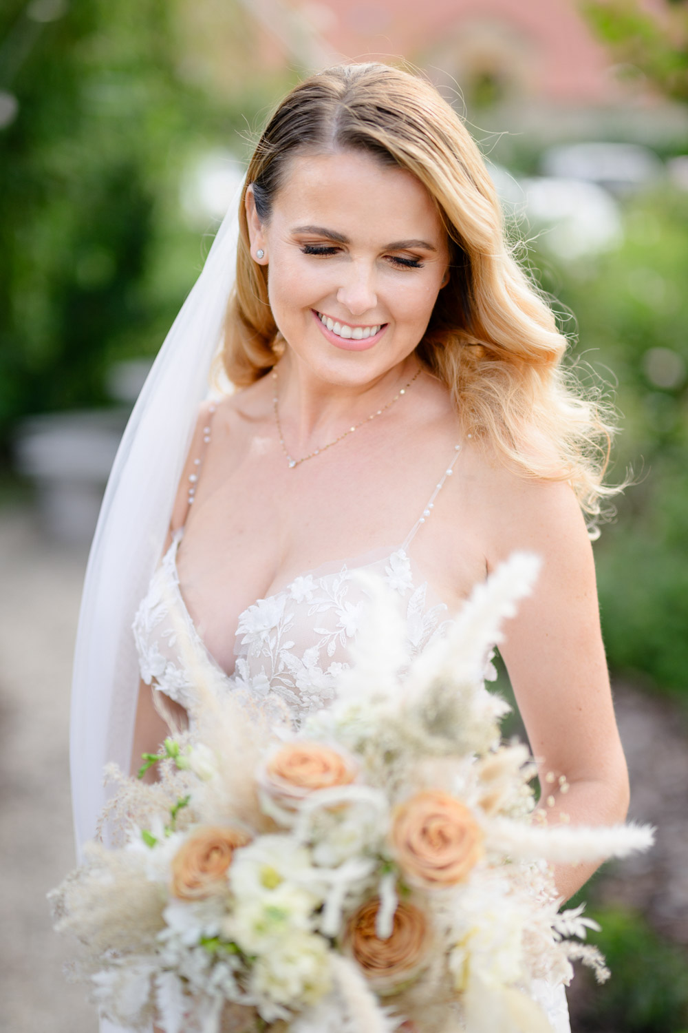 A bride in a beautiful white lace wedding dress holding a bouquet of peach and white flowers, standing outdoors in garden at Na Kmine