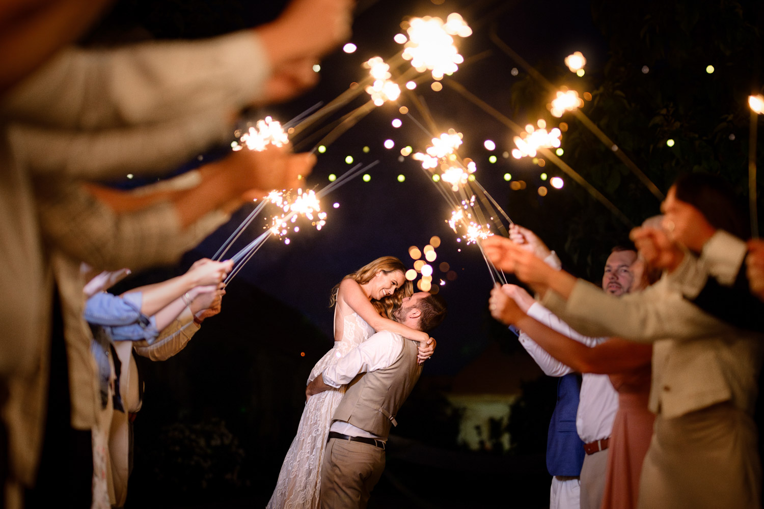A newlywed couple being lifted in the air, surrounded by friends and family holding sparklers, celebrating their wedding night at wedding venue Na Kmine.