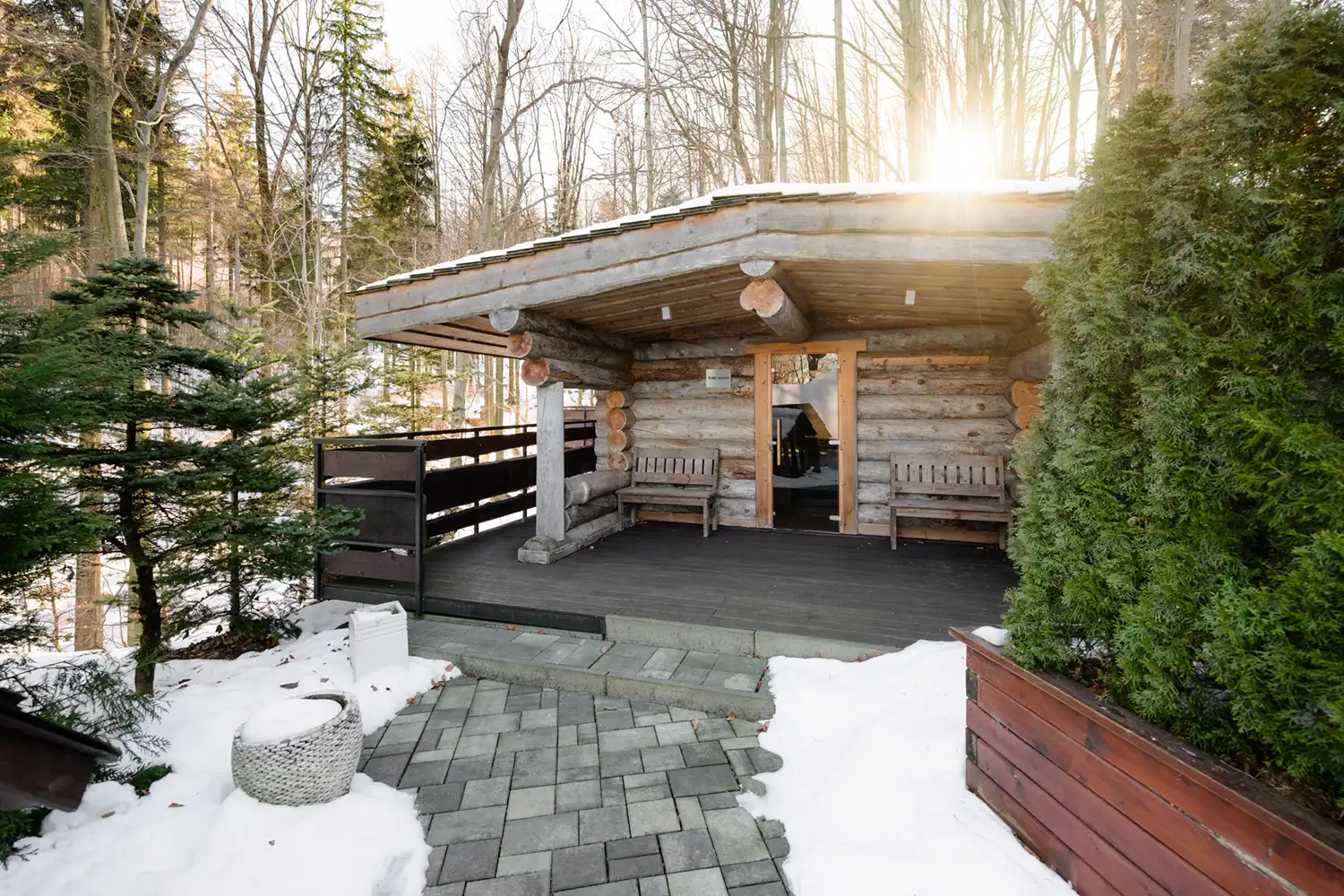 Exteriér dřevěného srubu s privátní kelo saunou v resortu Cattaleya, zasazený do zasněžené lesní krajiny s terasou a prosvítajícími slunečními paprsky.
