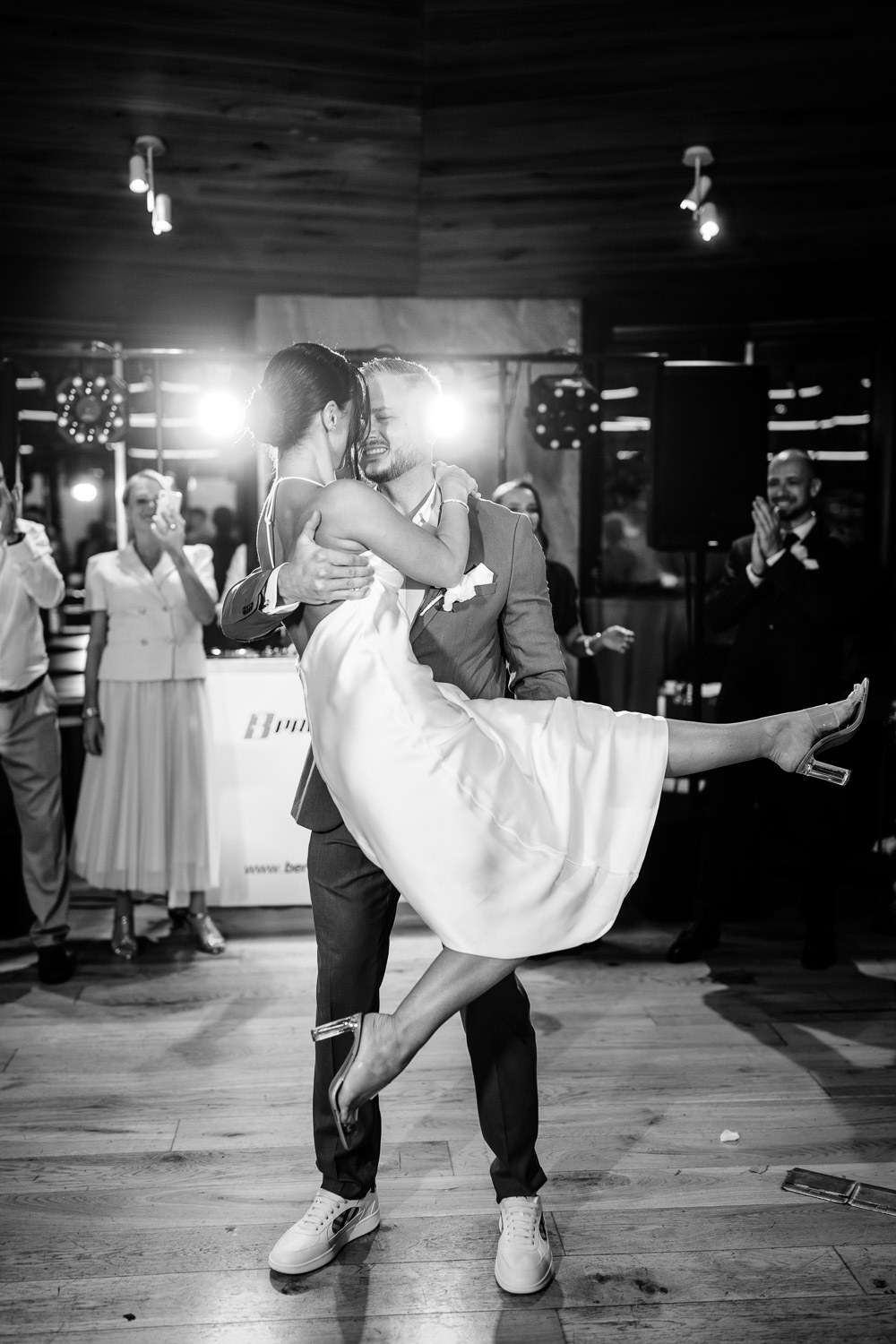 První tanec novomanželů: ženich zvedá nevěstu ve svatebních šatech, černobílá atmosféra plná radosti a oslavy na svatbě v elegantním sále.