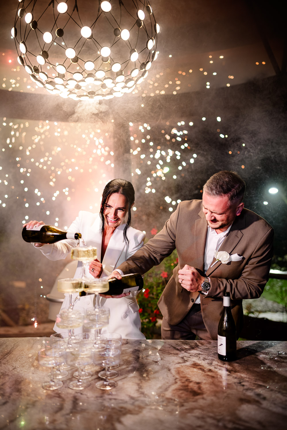 Wedding celebration: newlyweds pour champagne together from a tower of glasses, romantic atmosphere with lighting and fireworks at the Cattalaya resort.