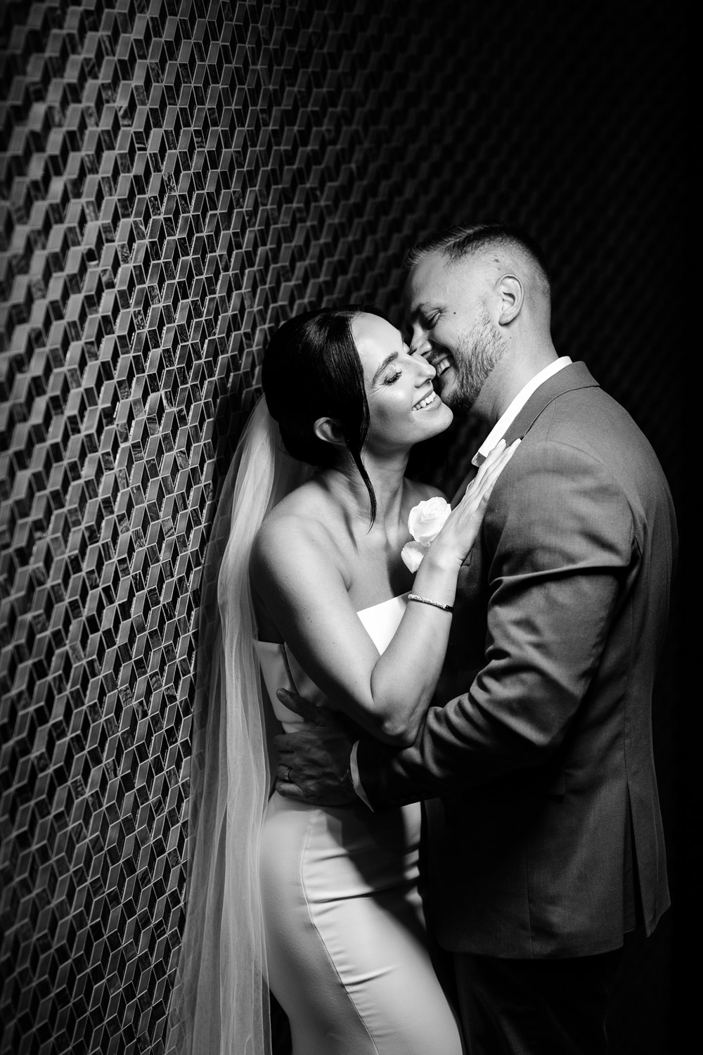 Black-and-white wedding portrait: newlyweds embracing in front of a modern geometric wall, the bride wearing a veil and the groom in an elegant suit