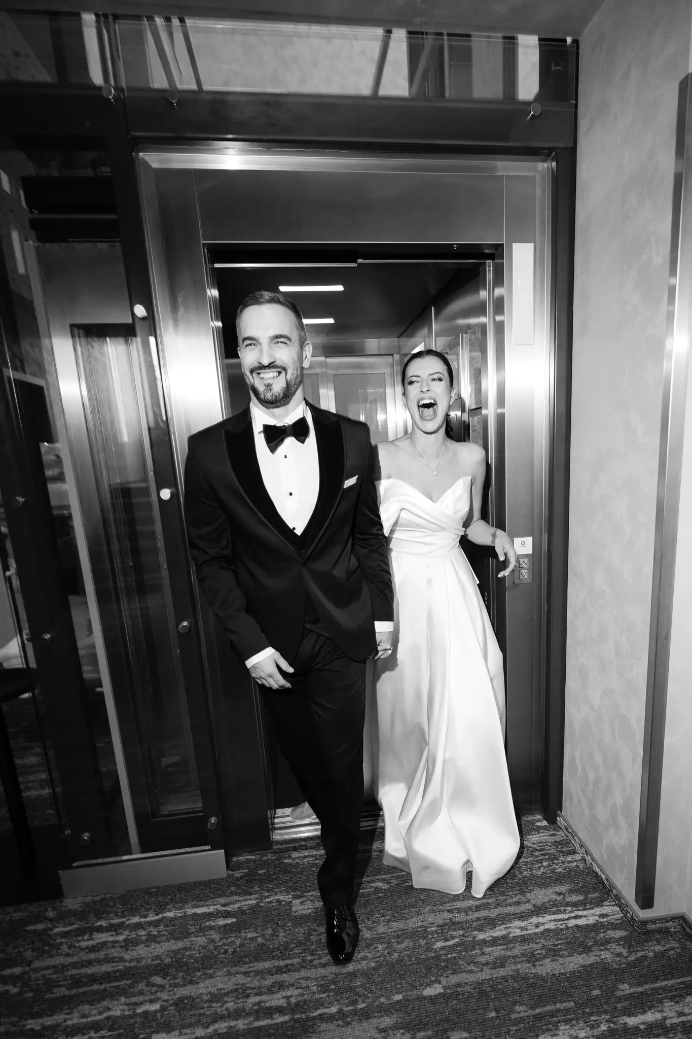 B&W photo of a laughing couple in an elevator. Minimalist dress, tuxedo. Wedding editorial Hotel Passage Brno. By: Jindrich Nejedly