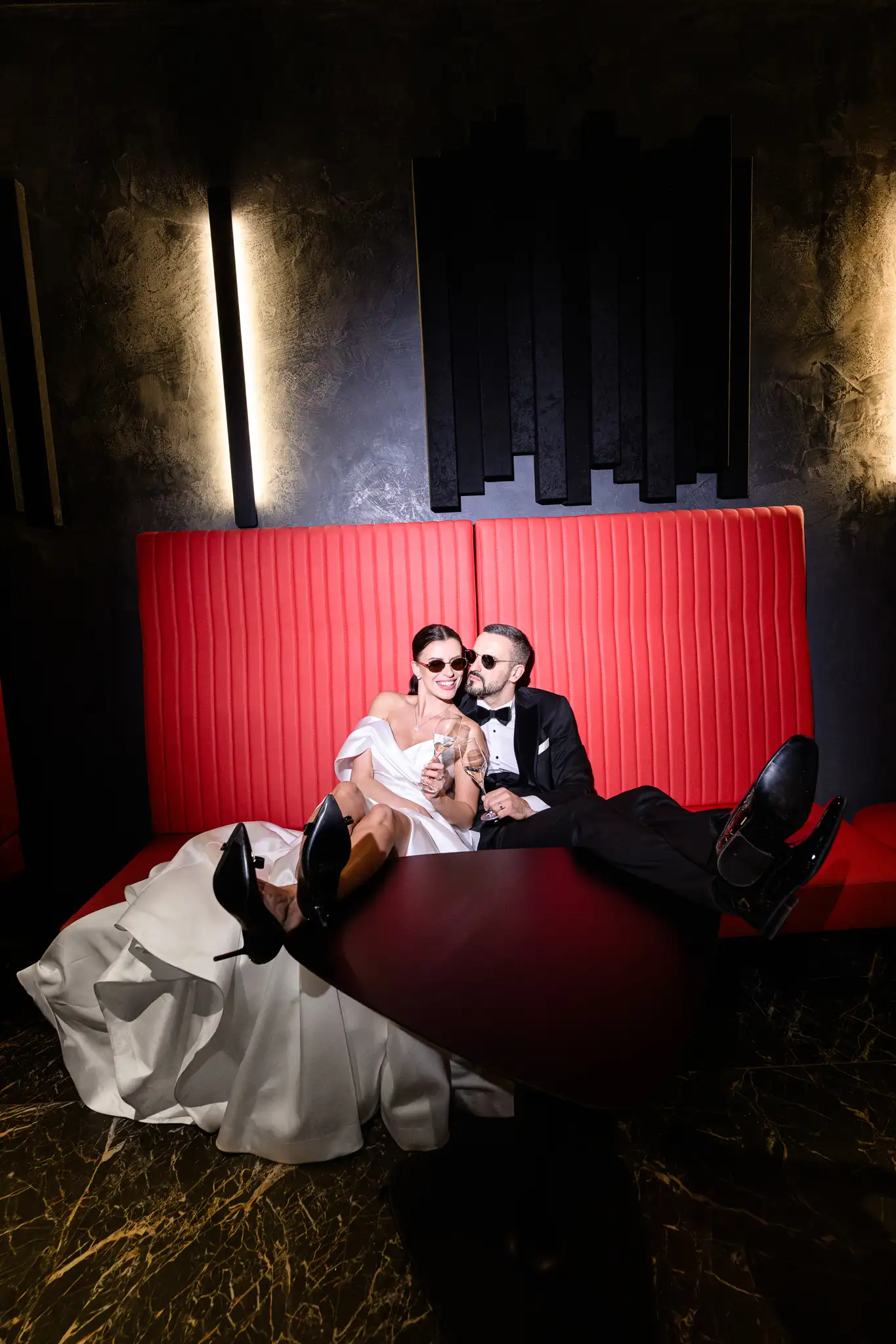 Wide-angle photo of newlyweds with feet on the table on a red booth. Rebellion and luxury. Hotel Passage Brno. By: Jindrich Nejedly