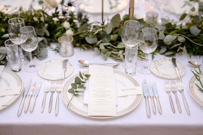 A luxurious wedding table setting with silver cutlery, menus on the plates, and a sprig of eucalyptus in Augustine hotel.