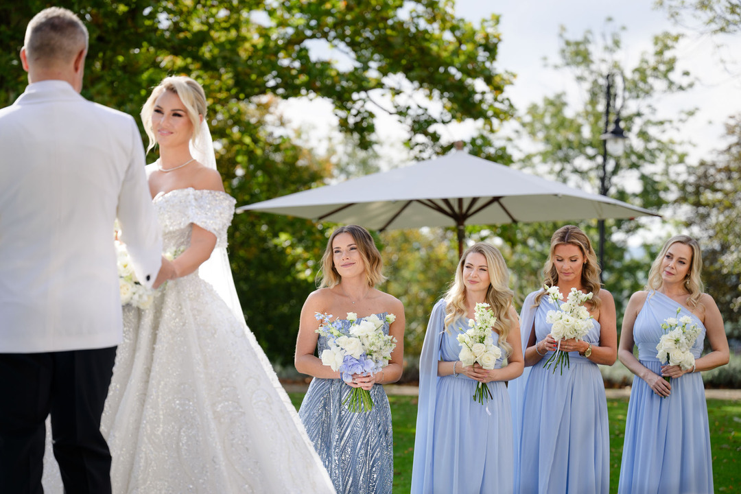 Bride and groom standing together during an elegant outdoor wedding ceremony at Chateau Mcely, with bridesmaids in light blue dresses holding bouquets.