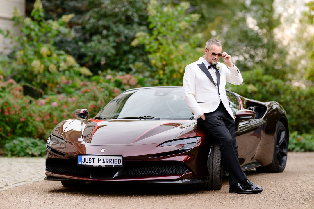 Stylish groom in a white tuxedo leans on a luxury Ferrari with a "Just Married" license plate at his Chateau Mcely wedding.