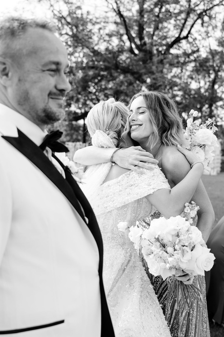 A heartfelt moment as the bride hugs a maid of honor, with the groom smiling nearby, capturing the emotion of a chateau wedding.