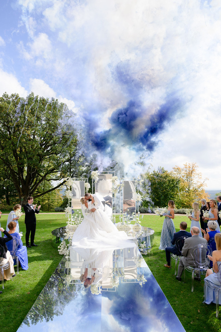 Newlyweds kissing under a floral arch with blue smoke in the background, reflecting in a mirrored aisle at a chateau wedding.