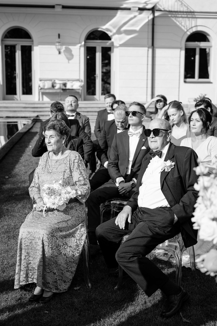 Guests seated outdoors during a wedding ceremony at Chateau Mcely, with elegant attire and a chateau building in the background.