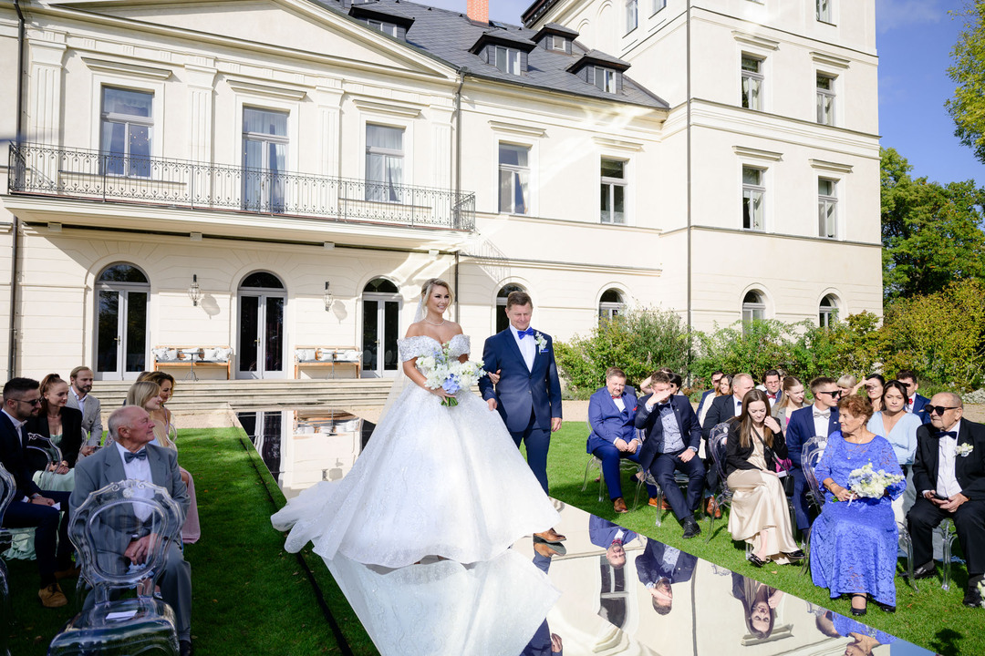 Nevěsta doprovázená svým otcem jde na svatební obřad na luxusní svatbě v Chateau Mcely, obklopeni elegantně oděnými hosty.