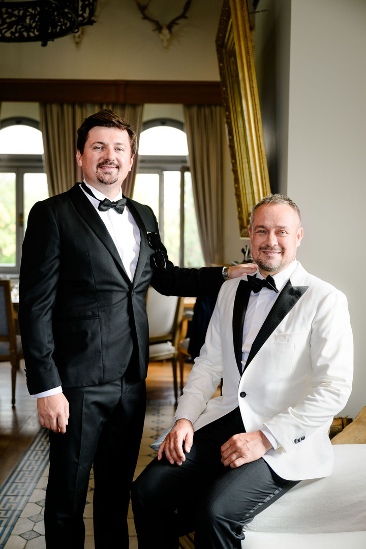 Groom in a white tuxedo and best man in a black tuxedo, posing together at a luxurious chateau wedding.