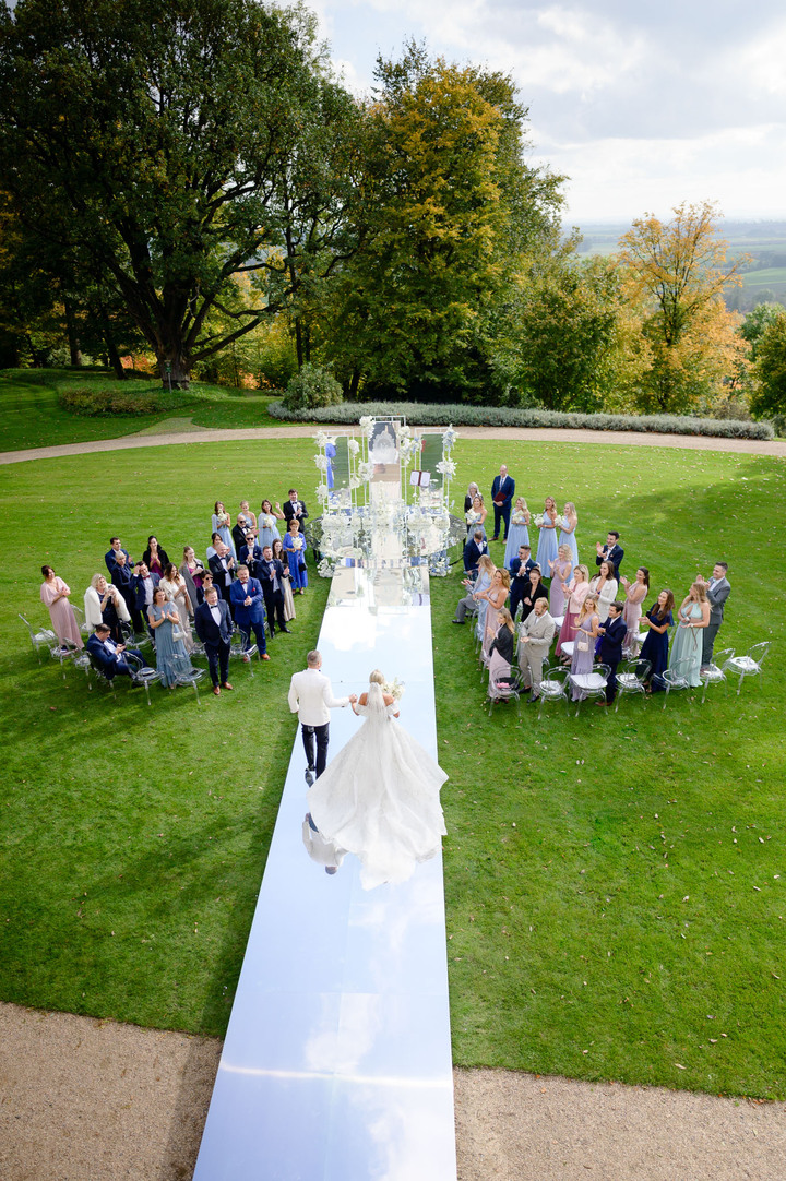 A bride and groom walking down a white aisle in a lush garden, surrounded by guests at a luxury chateau wedding.