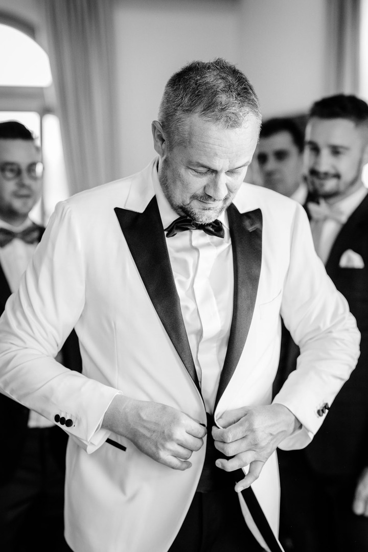 A groom adjusting his white tuxedo with black lapels, surrounded by groomsmen.