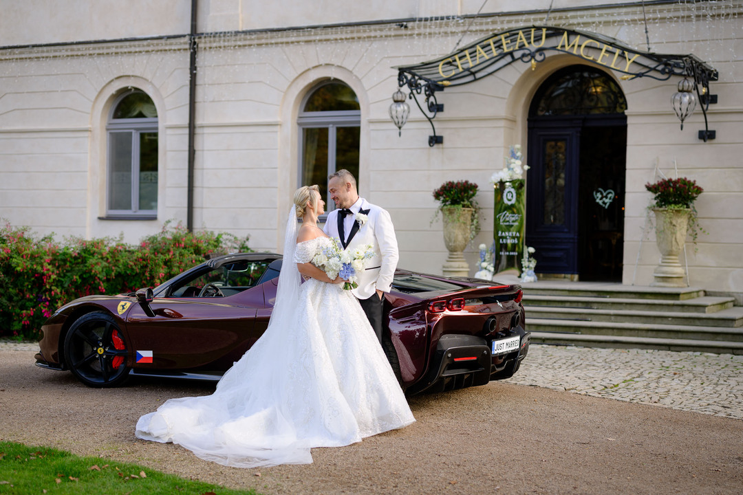 Nevěsta a ženich stojí před zámkem Mcely s luxusním sportovním autem, zdůrazňují eleganci svého svatebního dne.