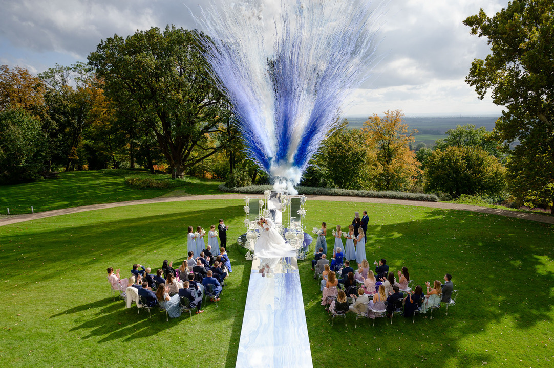 Outdoor wedding ceremony at Chateau Mcely with guests seated on a lush green lawn, a mirror aisle, and a dramatic blue and white confetti display.