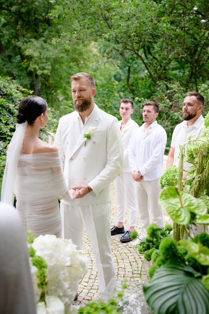Debbie Brown's wedding in Karlovy Vary: The fashion designer and the groom exchange vows in the garden of Villa Julius & Emma P. Witnesses in white attire observe the romantic ceremony, surrounded by greenery.
