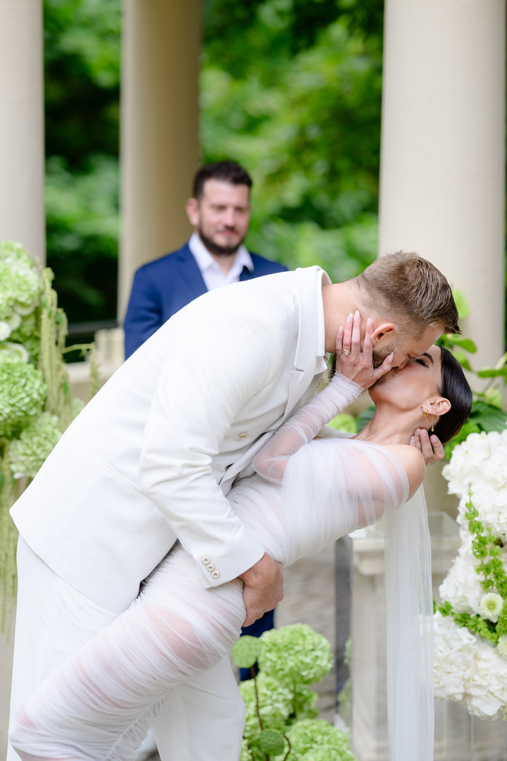 Bride Debbie Brown and the groom share a passionate kiss during the ceremony, surrounded by floral decorations with the officiant in the background.

