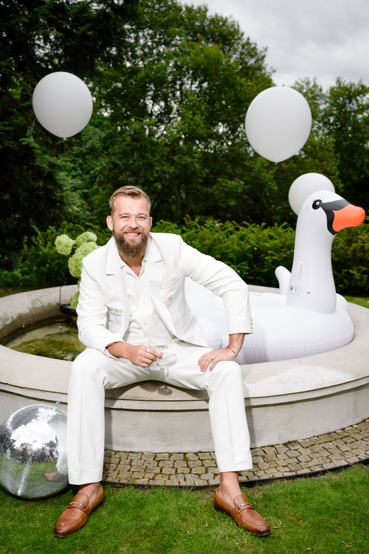 The groom in a white suit sits by the fountain on an inflatable swan, surrounded by balloons and a disco ball.
