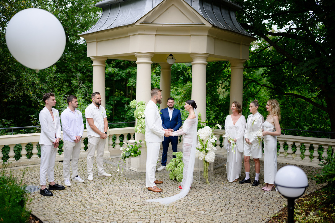 The ceremony in the gazebo at Villa Julius & Emma P. – in the center, the bride and groom hold hands, with friends on the right and left. Everyone is dressed in white.
