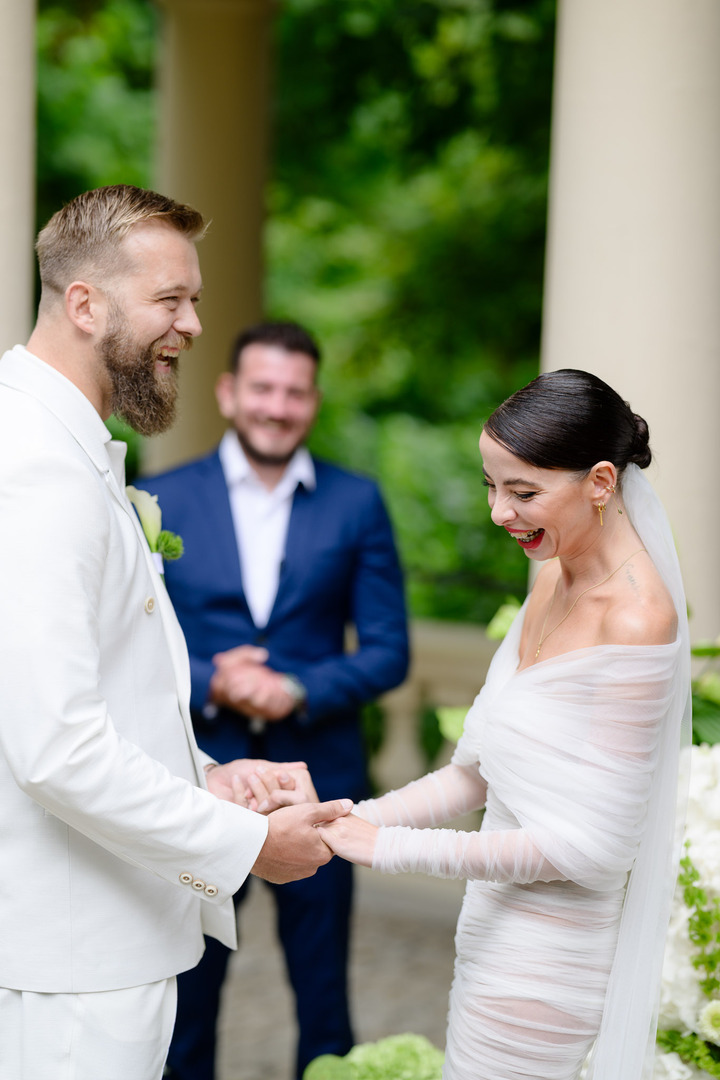 Fashion designer Debbie Brown, with a joyful smile, exchanges rings with the groom in the garden of Villa Julius & Emma P., while the witness observes the emotional moment.
