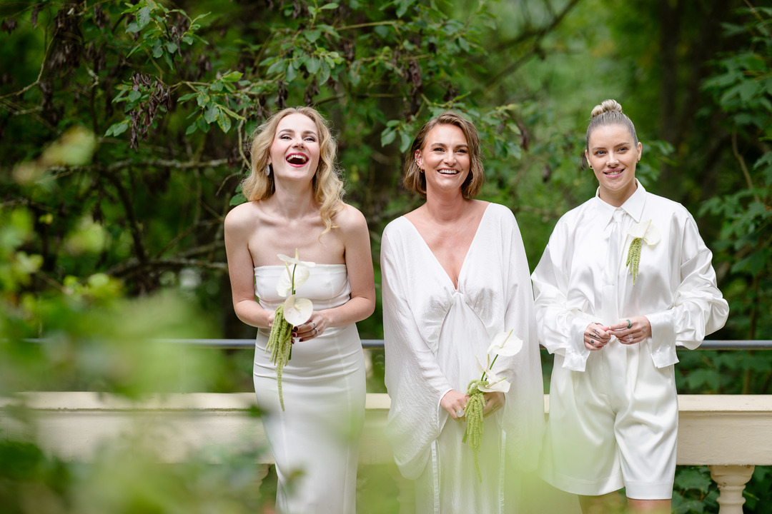 Three bridesmaids in white dresses, waiting for the bride, laughing and holding white lilies.
