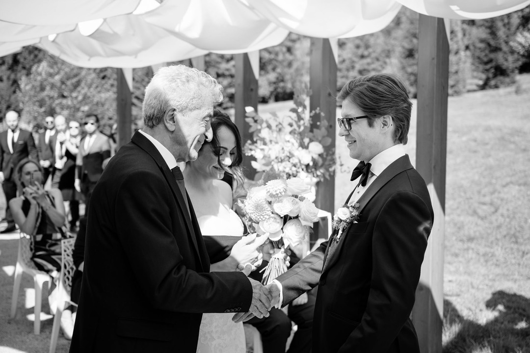 The bride’s father and the groom share a friendly clasp of hands.
