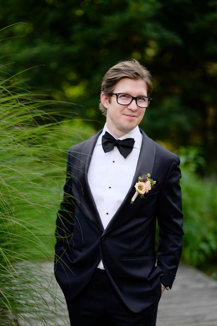Groom in a classic black tuxedo with bow tie and boutonnière, standing outdoors in the green nature of Beskydy Mountains.