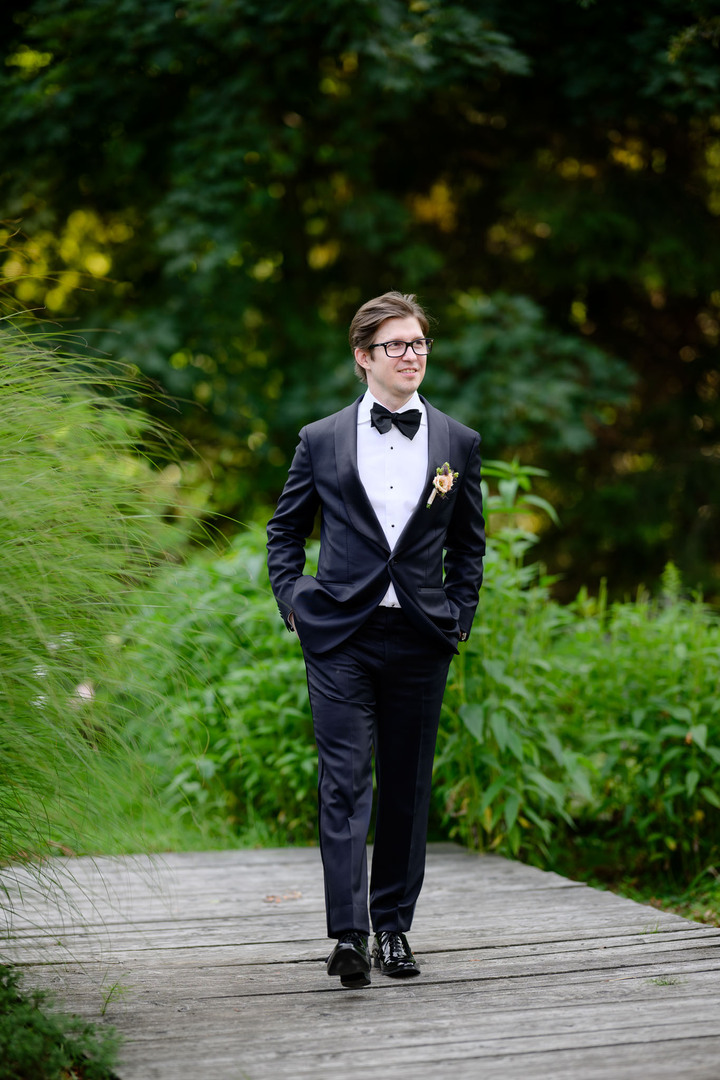 Groom in a classic black tuxedo with bow tie and boutonnière, standing outdoors in the green nature of Beskydy Mountains, Jindřich Nejedlý