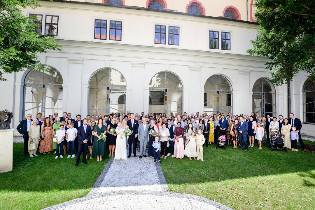 Společné foto všech svatebčanů před prosklenou stěnou hotelu Augustine. Ženich s nevěstou stojí uprostřed, nevěsta drží na rukách malou dcerku.