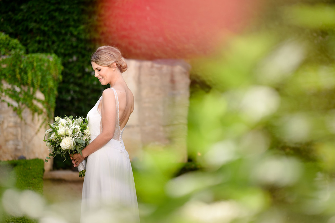 Pohled skrz květiny na nevěstu, která k nám stojí zády, v ruce drží svatební květinu a jemně se usmívá.
