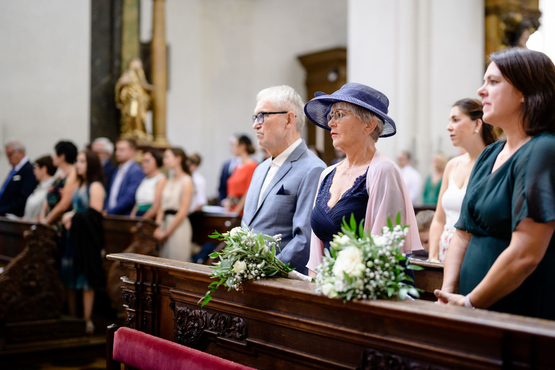 Záběr na rodiče nevěsty v kostelních lavicích, stojí a dojatě pozorují obřad.