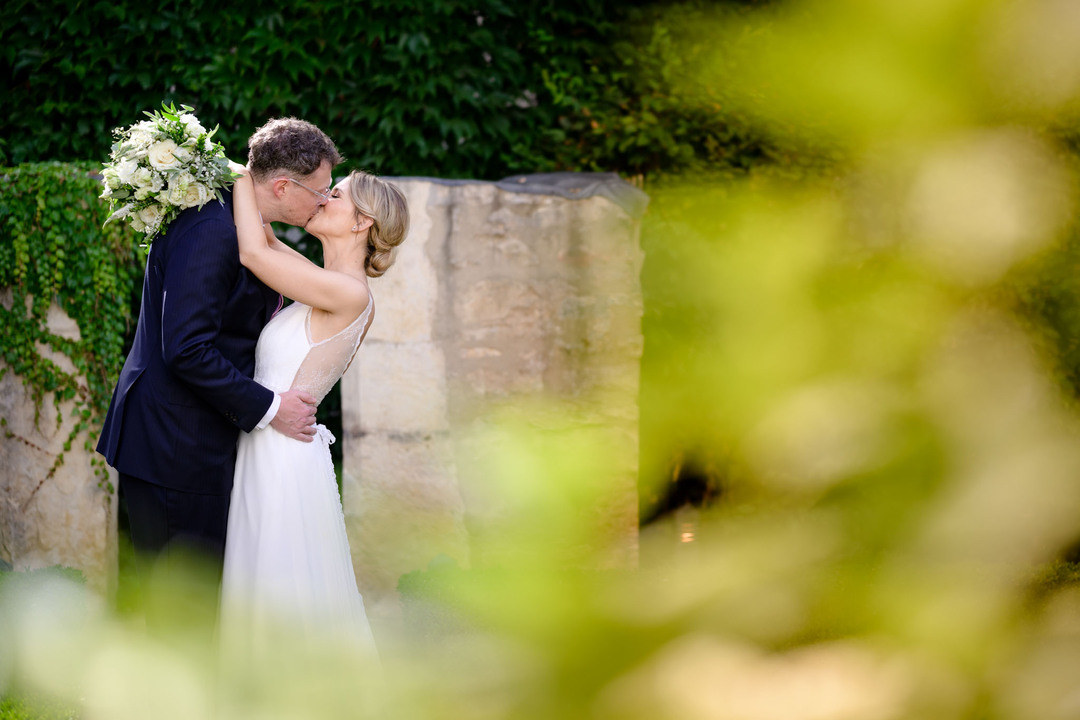 Nevěsta a ženich se líbají v zahradách hotelu Augustine v Praze, ženich drží nevěstu za boky, fotografie je focená zpoza zeleně.