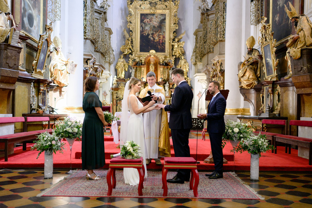 Záběr na pětici lidí - uprostřed farář, nevěsta se snubním prstenem, ženich, který nastavuje  ruku a po stranách svědek a svědkyně.