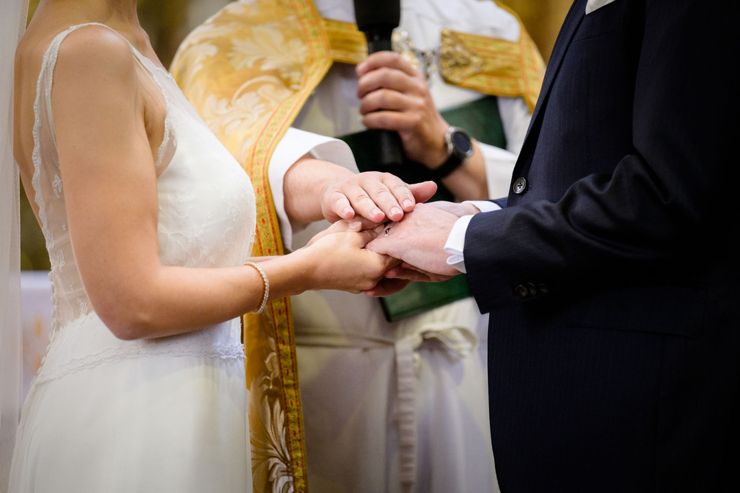 Pár se drží při obřadu za ruce, farář má svou ruku položenou na jejich a mluví do mikrofonu.