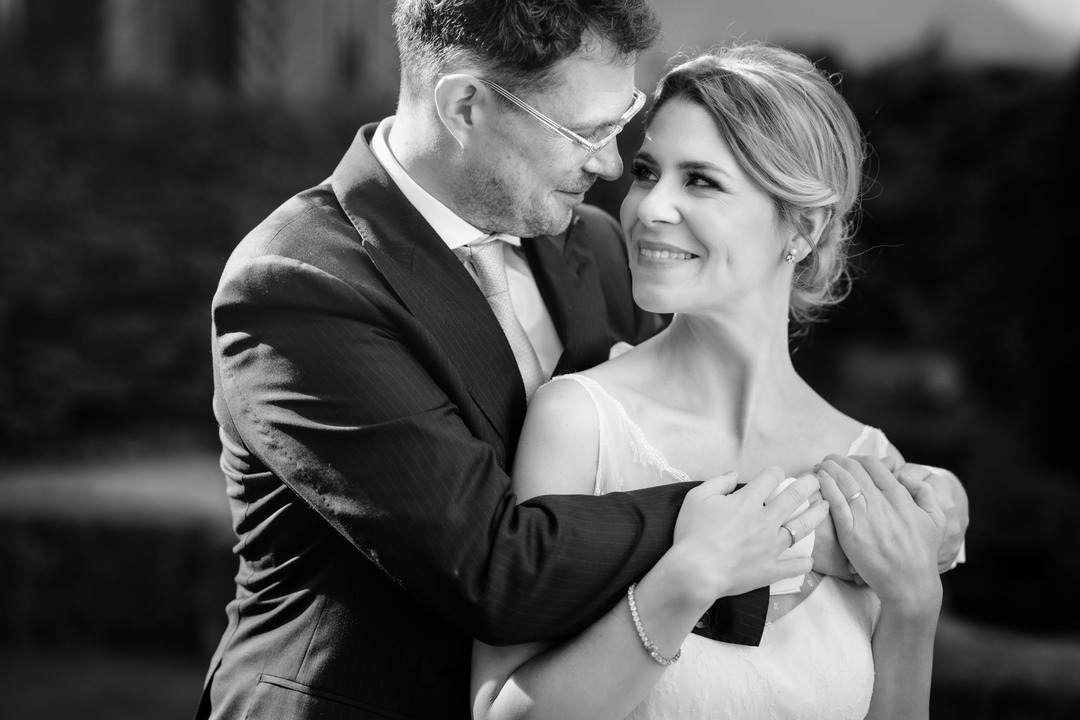 Černobílá fotografie - ženich zezadu objímá nevěstu, hlavy mají těsně u sebe, dívají se vzájemně do očí a usmívají se na sebe.