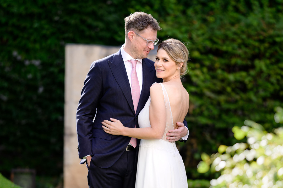 Nevěsta drží ženicha za boky, je k němu přitisknutá, stojí zády k nám, ale dívá se do fotoaparátu. Ženich se s láskyplným úsměvem dívá na svou ženu.