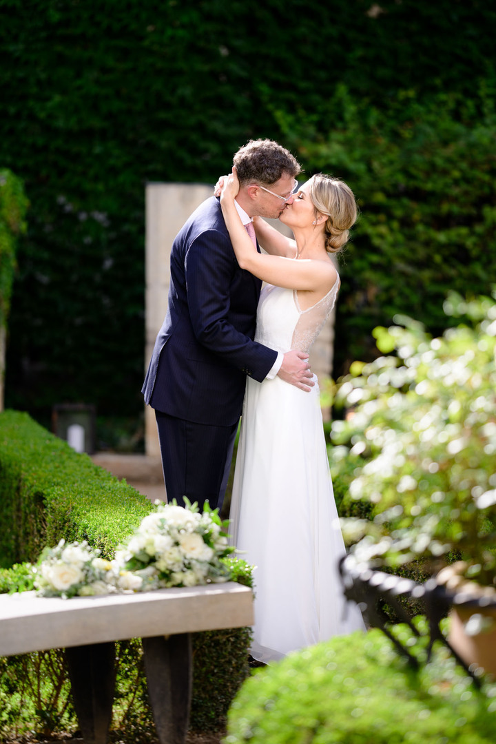 Novomanželé stojí v zahradě hotelu Augustine, ženich drží nevěstu za boky, nevěsta drží ženicha kolem krku, líbají se.