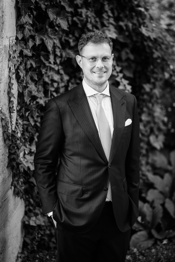 Čb záběr na ženicha - stojí s rukama v kapsách a usmívá se, za ním je stěna pokrytá břečťanem. Zblízka jde vidět, že má jeho oblek jemné proužky.