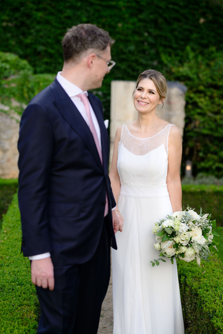 Novomanželé kráčí zahradou u hotelu Augustine, drží se za ruce, ženich jde vpředu a ohlíží se na nevěstu, ta se na něj usmívá.