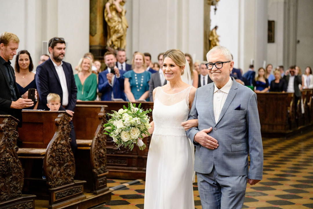 Nevěsta prochází uličkou v kostele spolu se svým otcem, ona má bílé šaty s krajkou na hrudi, on má šedý oblek a nosí černé brýle. Oba se usmívají.